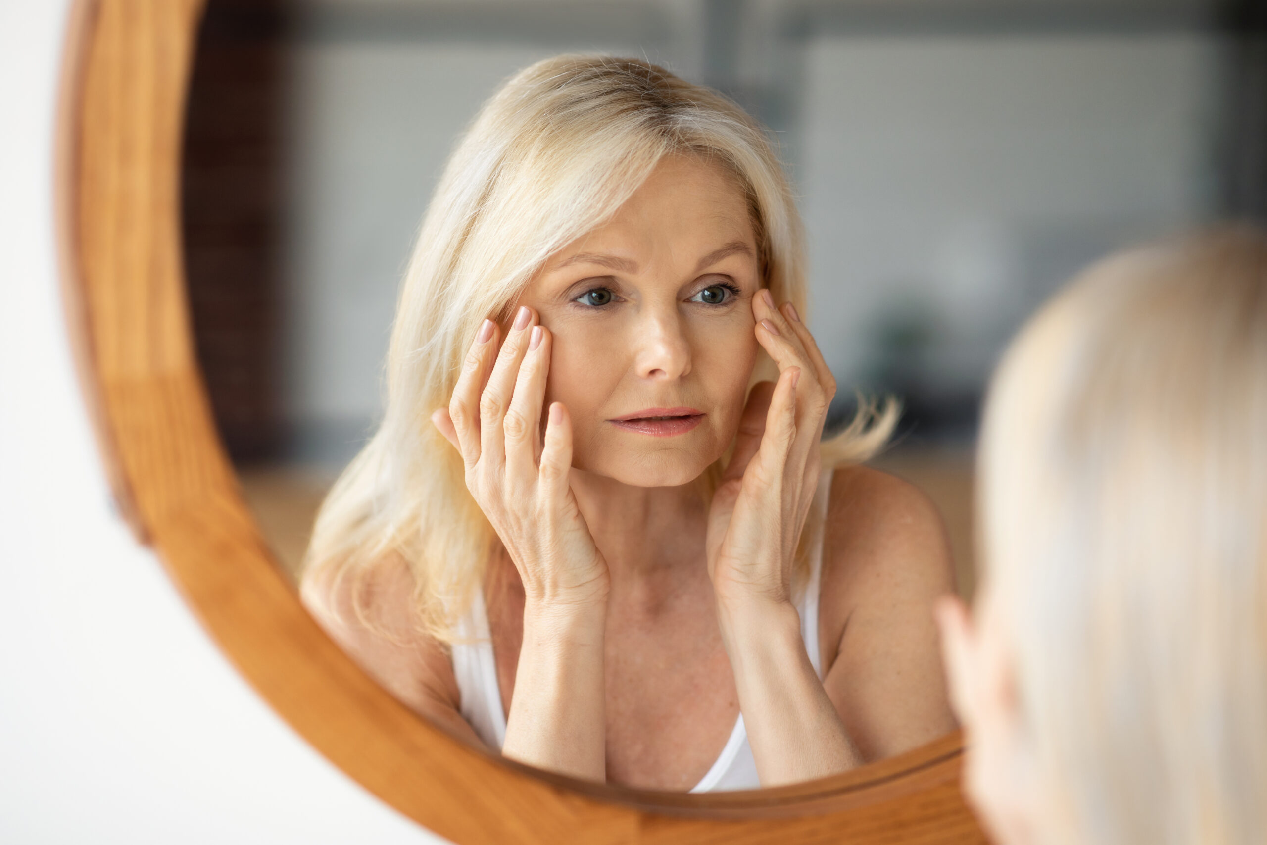 Aged skin problems. Upset senior woman looking at mirror and touching face, lady noticed wrinkles near eyes